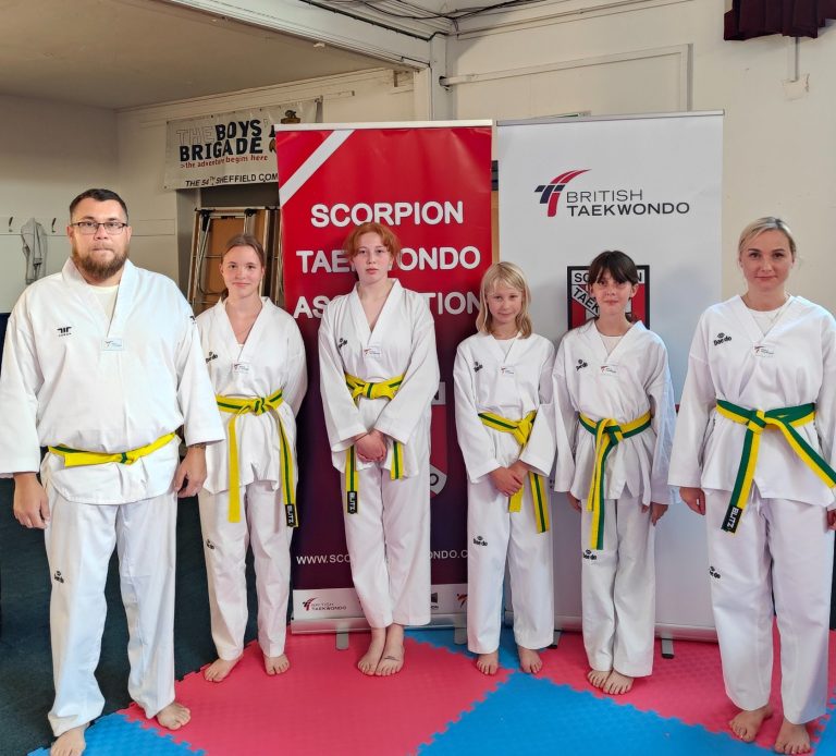A group of students stand in front of the Scorpion logo wearing white martial arts suits and yellow belts
