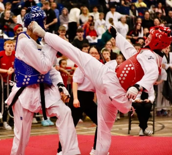 A taekwondo competitor wearing red protective gear executes a high roundhouse kick to the head of an opponent in blue gear during a match.