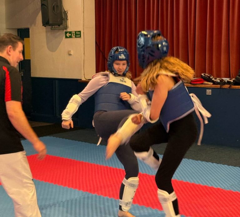 Two teenage girls are sparring with their coach looking on and supporting. Both girls are wearing protective gear